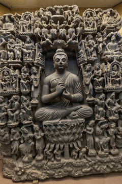View of an intricate Gandhara art stone sculpture depicting Buddha seated in a meditative pose on a lotus throne, surrounded by smaller figures and ornate carvings in Lahore, Pakistan.