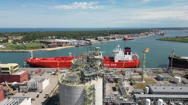 Aerial drone view of red gas tanker ship docked at Ravenna port near chemical refinery plant with gas flare stack burning bright on sunny day in Emilia Romagna Italy 