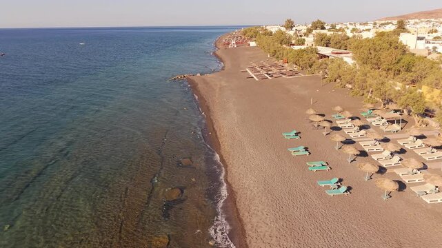 Drone shot of calm volcanic black sand beach with sunbeds along Aegean coast in Perissa Santorini Greece
