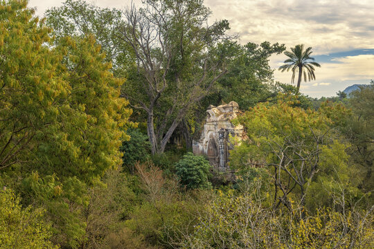 View of the historic Malkana Temple nestled among lush green trees and a palm tree under a cloudy sky in Dalwal, Punjab, Pakistan.