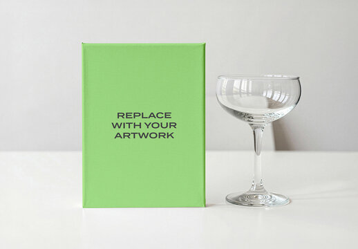 Vertical canvas mockup with green textured surface positioned beside a clear glass coupe on white tabletop, minimalist studio scene for artwork or branding display, template print digital design