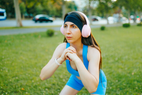 Beautiful young woman with headphones doing a fitness squat workout in the summer city.