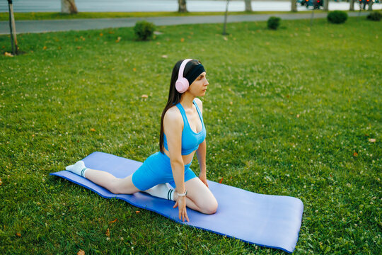 Fit young woman with headphones stretching on a purple yoga mat in the green park.