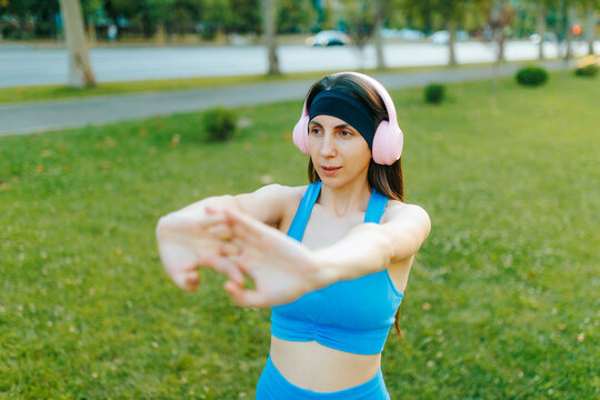 Active woman listening to music performing an arm stretch routine in the sunny summer park.