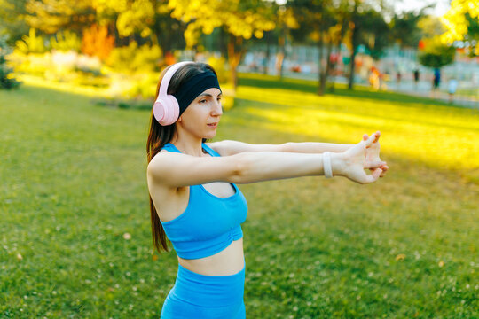 Active fit woman listening to music and doing an arm stretch exercise in the sunny park.