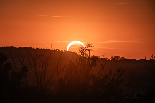 View of a partial solar eclipse during sunset with silhouettes of trees and wind turbines on the horizon in Piekary Slaskie, Silesian Voivodeship, Poland.