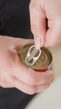Close-up top view shows hands opening pull-tab can, lifting ring, folding tab back and puncturing lid while fingers steady metal container; visible manual action, skin detail, reflective surface and
