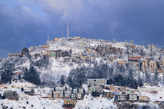 View of the snow-covered hillside town with buildings nestled among frost-laden trees under a dramatic overcast sky in Murree, Punjab, Pakistan.