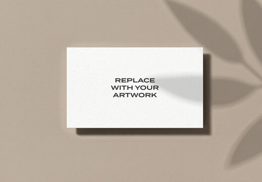 Textured business card mockup on a beige wall with leaf shadow, minimalist stationery branding presentation, print design template, vertical flat lay, clean office desk display, paper texture
