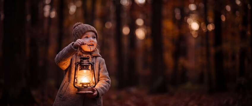 Little Girl Holding Glowing Vintage Oil Lantern in Dark Autumn Forest, Atmospheric Cinematic Stock Photo