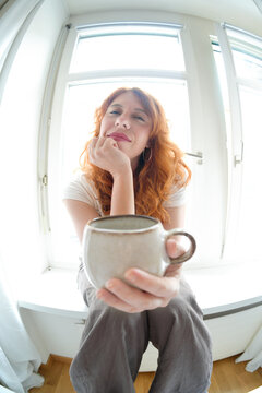Redhead woman offer coffee mug in bright home interior