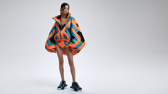 Modell in oversized Streetwear-Jacke mit orangeblauem Camouflage-Muster und Shorts, wei&szlig;er Studiohintergrund, extravagantes Modeshooting.