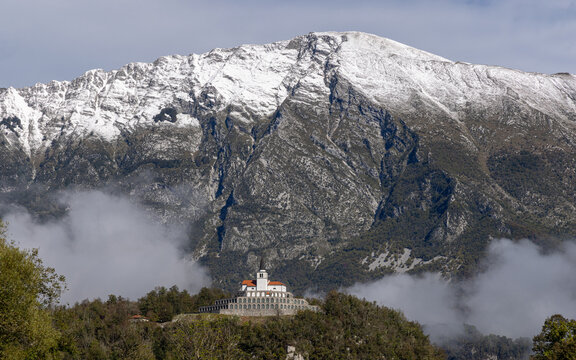 View of Javorca Memorial Church perched on a hill with snow-capped mountains in the background and low-lying clouds in Tolmin, Slovenia.