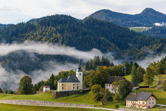 View of a historic church with a tall steeple nestled in lush green hills, surrounded by morning mist and dense pine forests, Skofja Loka, Slovenia.