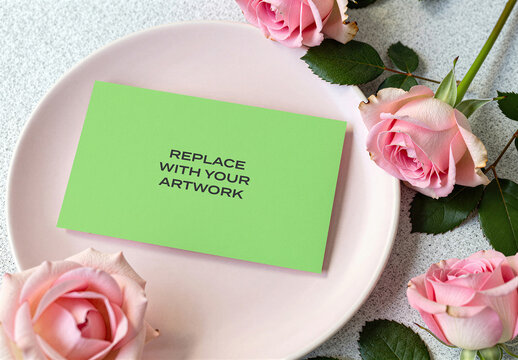 Business card mockup on a pink plate surrounded by roses and green leaves, flat lay composition with floral arrangement, realistic stationery branding presentation, vertical rectangular surface