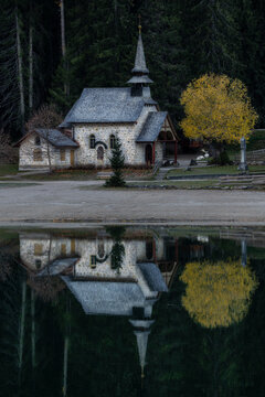 View of the stone church reflecting in the calm lake, surrounded by forest and a yellow tree in the Dolomites, Campitello di Fassa, Trentino-Alto Adige, Italy.