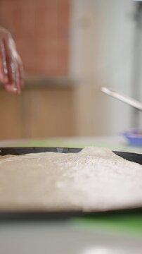 Close-up of hand pressing and smoothing raw dough on dark rimmed shallow tray, showing steady forward palm motion, soft ridges, finger marks, and tactile texture on lightly dusted surface