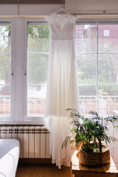 Bridal wedding dress hanging by sunlit window indoors