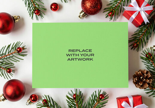 Horizontal card mockup with Christmas ornaments and pine branches arranged around it on a white surface for holiday stationery presentation, flat lay top view, product copy space