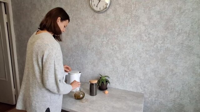 Woman preparing tea in a kitchen while checking the clock on the wall in a home setting