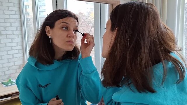 Woman applying makeup while looking in a mirror in a bright indoor setting in the morning