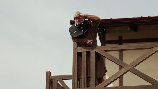 Older man in brown coat and flat cap peers through binoculars while standing on rustic cross braced wooden observation platform, watching distant horizon under soft overcast light beside weathered