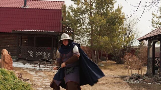 Helmeted reenactor in chainmail, cloak, and tunic lunges forward with raised sword while bracing shield in rustic homestead yard; extended arms, leaning stance, billowing cloak convey motion under