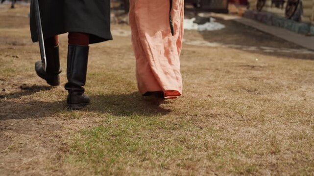Low view rear shot of two figures walking across dry, patchy grass, with visible forward stride, lifted heels, trailing cloak, and flowing gown brushing ground while disturbing turf under muted