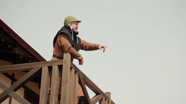Middle-aged man on elevated timber balcony under diffuse daylight, leaning over railing with gloved hand extended downward in focused inspection pose, wearing helmet, heavy coat, and work gloves