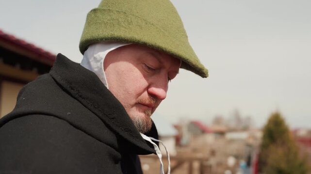 Close side profile of middleaged man wearing green knit cap with earflaps and heavy dark coat on elevated urban rooftop, facing right with focused neutral expression and cool outdoor light
