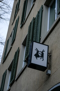 Zurich, Switzerland - March 15, 2026. Whimsical Dancing Rabbits Sign on Traditional Building Facade in Zurich. A low-angle shot captures a modern backlit square sign featuring a silhouette of two anth