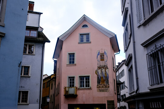 Zurich, Switzerland - March 15, 2026. Historic Pink Building with Medieval Fresco in Zurich Old Town. A low-angle shot captures a pastel pink residential building nestled between narrow alleys in the 