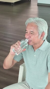 asian senior man sitting and drinking water from a plastic bottle in his living room, feeling refreshed and hydrated