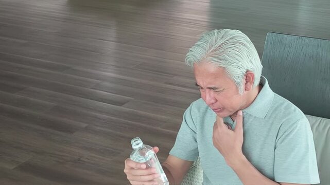 asian senior man choking while drinking water from a plastic bottle in his living room, medical emergency and distress
