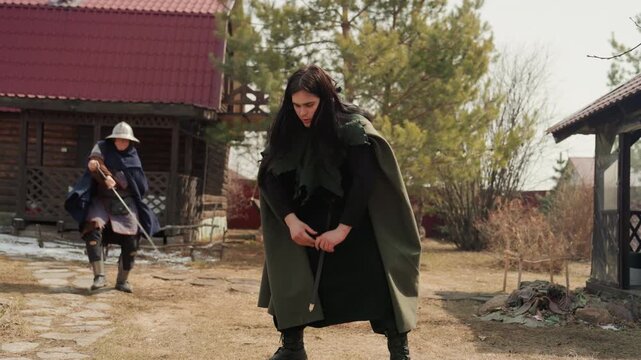 Woman in layered green cloak faces helmeted guard in dirt courtyard, cloak flowing with recent movement while she holds firm stance; guard steps back near spear, wooden buildings, grass, and muted