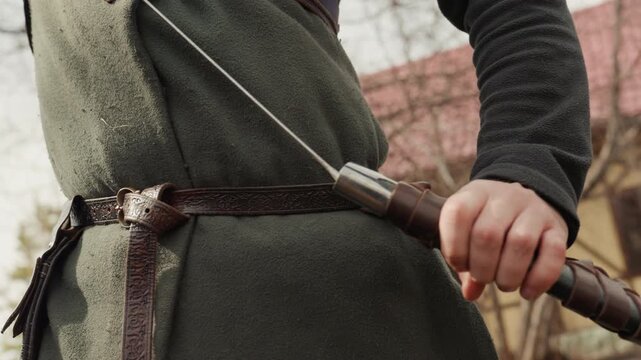 Tight closeup shows two hands gripping sword hilt and crossguard at hip level, with tense fingers, wrapped wrists, belt, scabbard, and muted outdoor light creating strong preparation mood