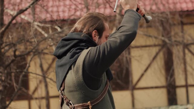 Archer pulls bowstring to full draw while companion lifts sword into powerful swing beside fence and tiled roof, with soft daylight, focused posture, visible strain, training energy, courtyard setting