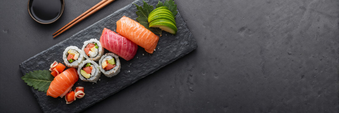 Assorted sushi and sashimi served on a slate plate with soy sauce, chopsticks, and avocado. Japanese cuisine concept with dark copy space for menu design and food promotion.