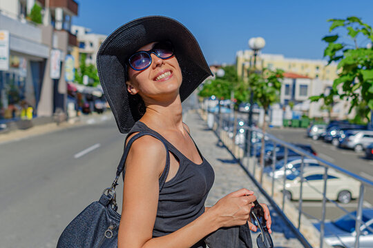 A smiling woman wearing a black hat and sunglasses enjoys a sunny day walking through a city street.
