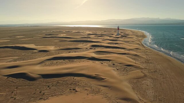Aerial drone flight over dramatic coastal sand dunes leading toward Punta del Fangar lighthouse on Mediterranean shoreline 