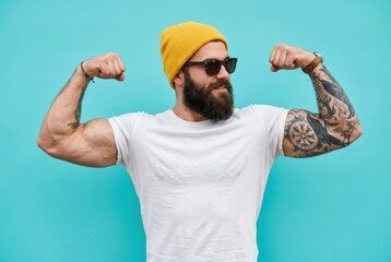 Naklejka premium Strong Muscular Bearded Man with Tattoos Flexing Biceps in White T-shirt and Yellow Beanie on Blue Background