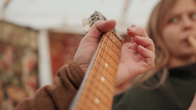 Tight low-angle view of left hand pressing strings on guitar neck, fingers forming chord changes while blurred companion remains in warm indoor background, creating focused musical mood