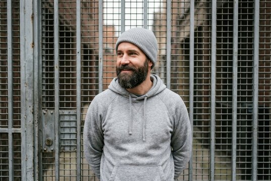 Portrait of a Mature Bearded Man in Grey Hoodie and Beanie Standing in Front of a Metal Gate in an Urban Setting