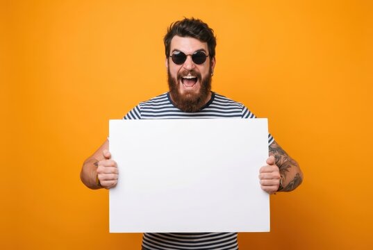 Excited Bearded Hipster Man in Sunglasses Holding Blank White Poster Board for Copy Space on Orange Background