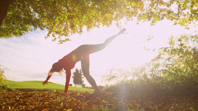 LENS FLARE, SLOW MOTION, DOF, LOW ANGLE VIEW: Backlit by bright autumn sunshine, a playful woman does a cartwheel under colorful tree. Energetic and carefree feeling in beautiful countryside setting.