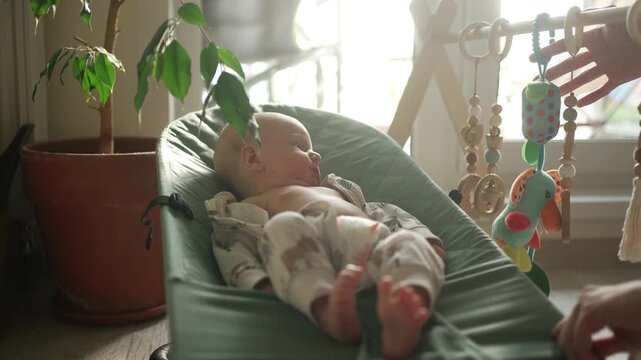 Newborn baby resting comfortably in a green bouncer while a mother's hand rocks the chair and interacts with the hanging toys, creating a warm and gentle domestic scene