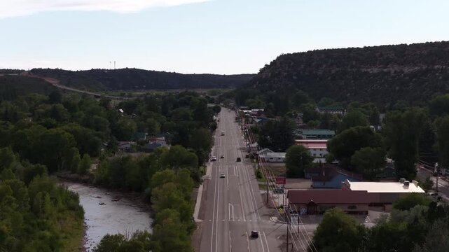 Aerial of small town street view in Dolores Colorado with Dolores River and forested hills with main street featuring commercial buildings in summer