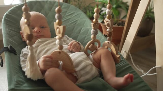 Curious newborn baby resting in a comfortable green bouncer, attentively observing and interacting with a hanging wooden mobile, promoting early sensory and motor skill development
