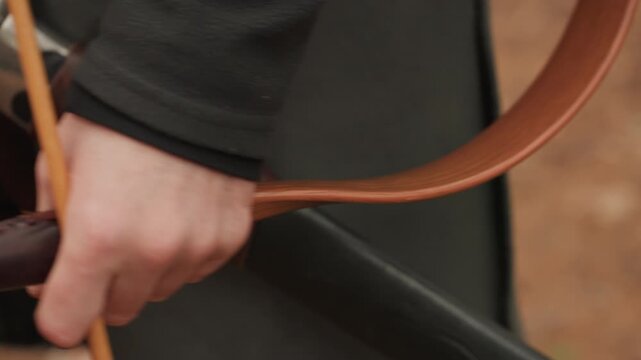 Close view of hand gripping bow riser and drawing string on wooden recurve bow, with nocked arrow, curved limbs under tension, steady finger placement, and focused outdoor detail in warm natural light