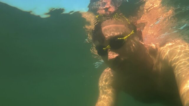 Man with goggles swimming submerged in murky green water, sunlight creating caustic patterns on his skin